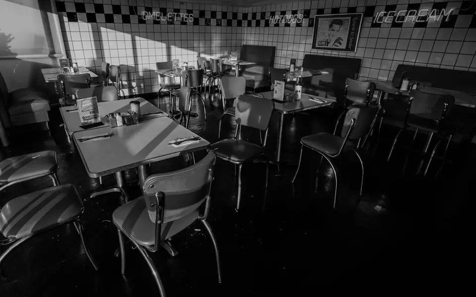 An empty diner with chrome chairs, small tables and a checkered border along the tiled wall.