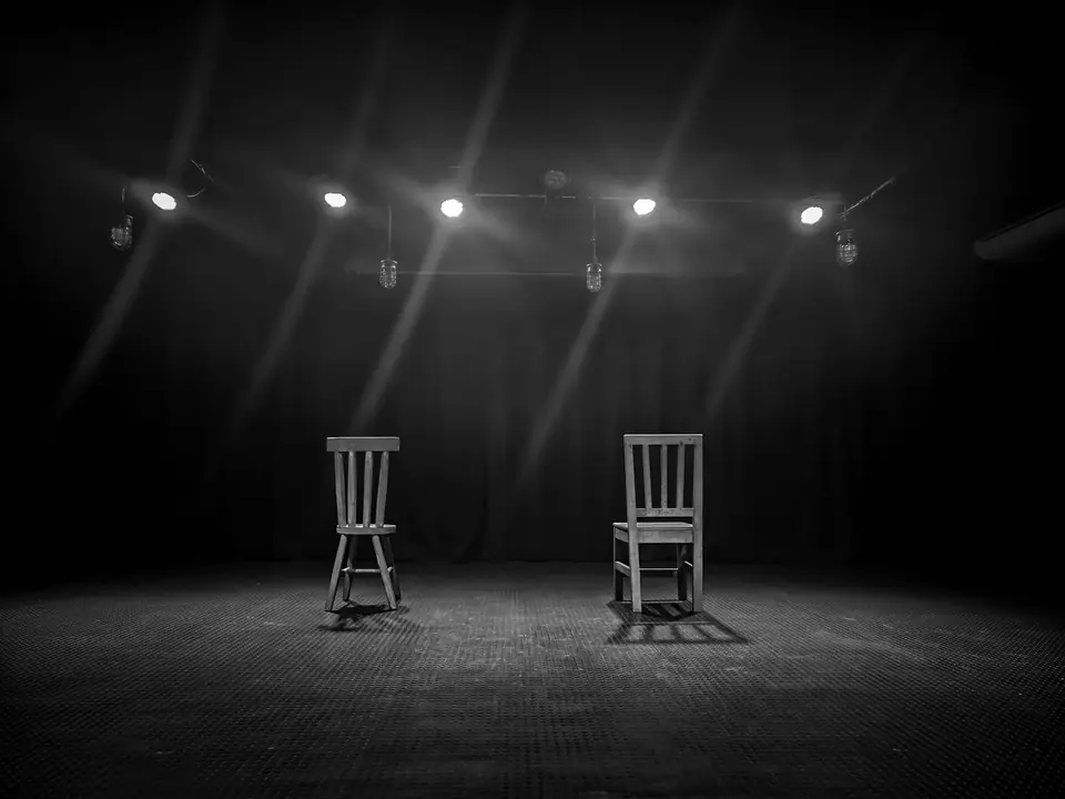 Two wooden chairs stand apart on an empty stage beneath a row of spotlights.