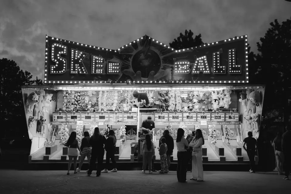 A group of people stands in front of a lit Skee-Ball booth with a marquee of bulbs at dusk.
