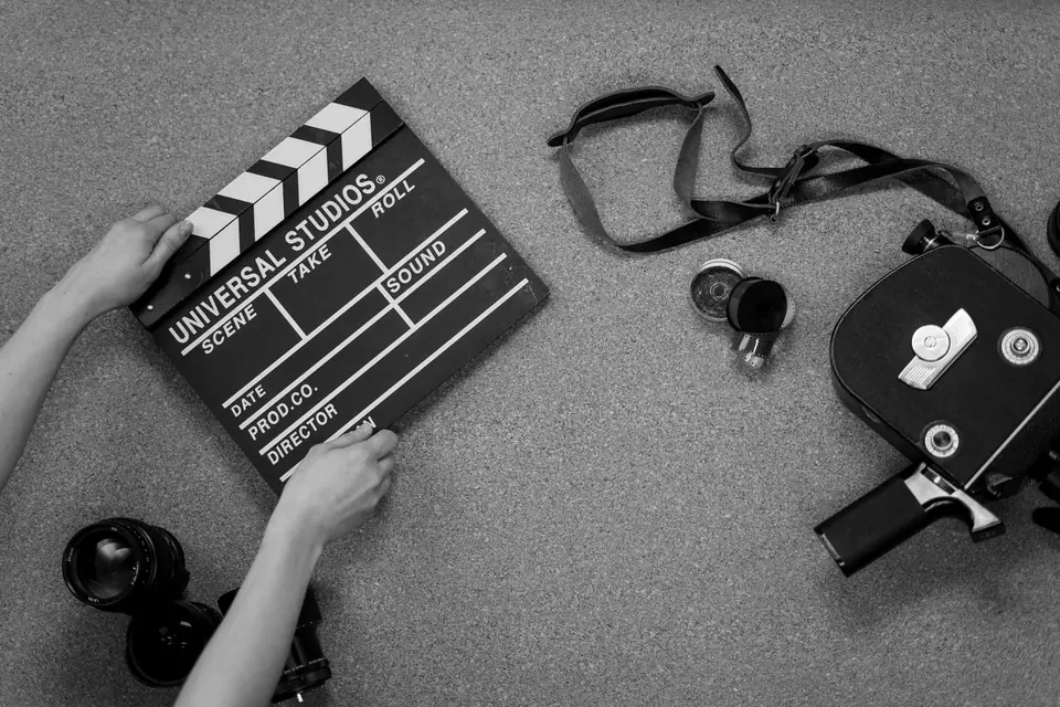 Hands hold a film clapperboard on the floor beside a vintage movie camera and lenses.