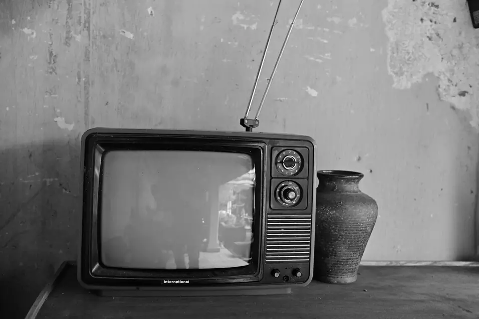 A small vintage television with an antenna and round dials sits beside a clay vase against a worn wall.