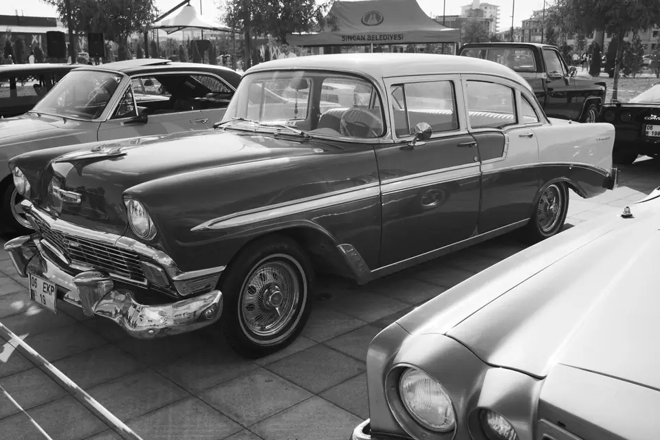 A two-tone 1950s sedan with chrome trim is parked among other classic cars on a paved lot.