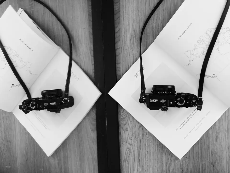 Two small black cameras with neck straps lie on open books placed on a wooden table.