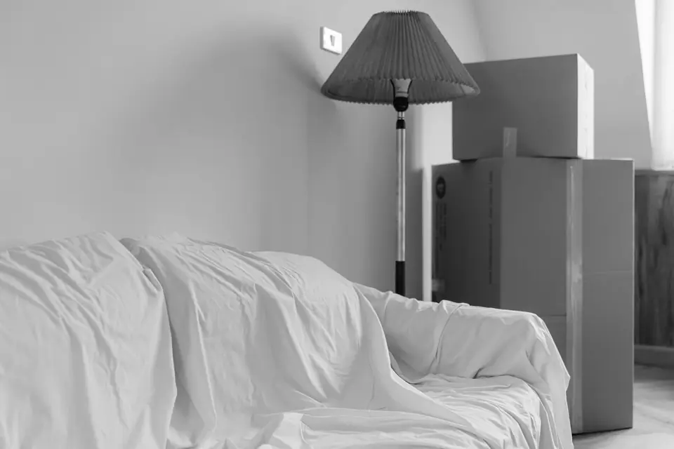 A sofa covered by a white sheet sits beside a floor lamp and stacked cardboard boxes in a plain room.
