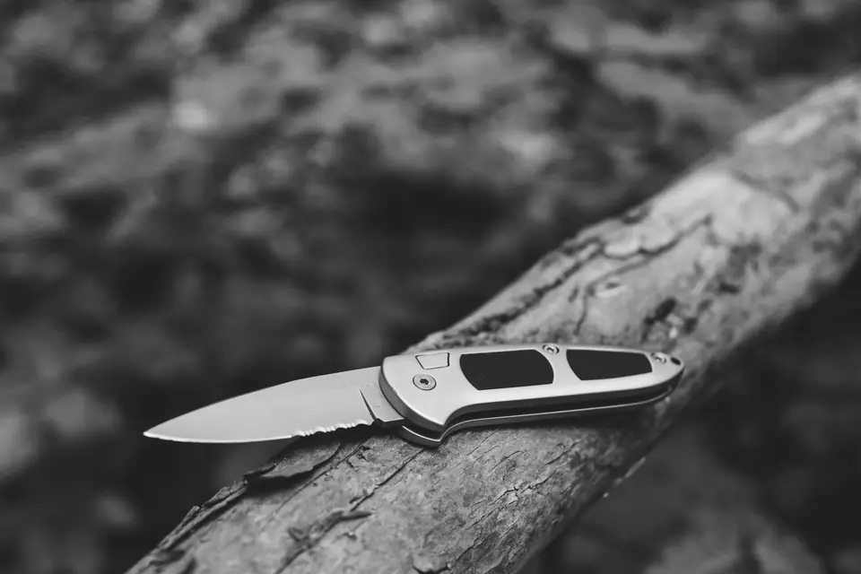 An open folding knife with a partly serrated blade rests on a rough tree branch outdoors.