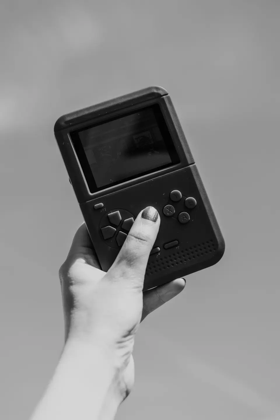 A hand holds up a small dark handheld game console with a screen and buttons against a bright sky.
