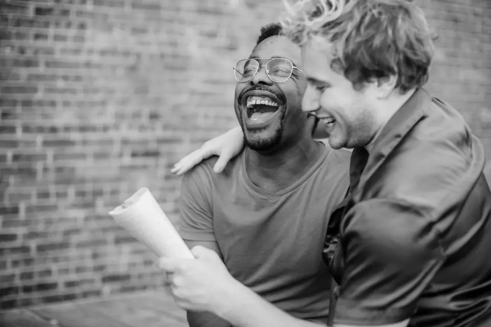 Two laughing men embrace in front of a brick wall, one holding a rolled paper.