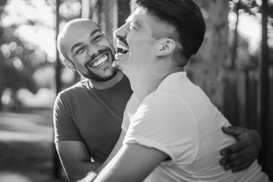 Two men laugh together outdoors, one with his arm around the other, trees blurred behind them.