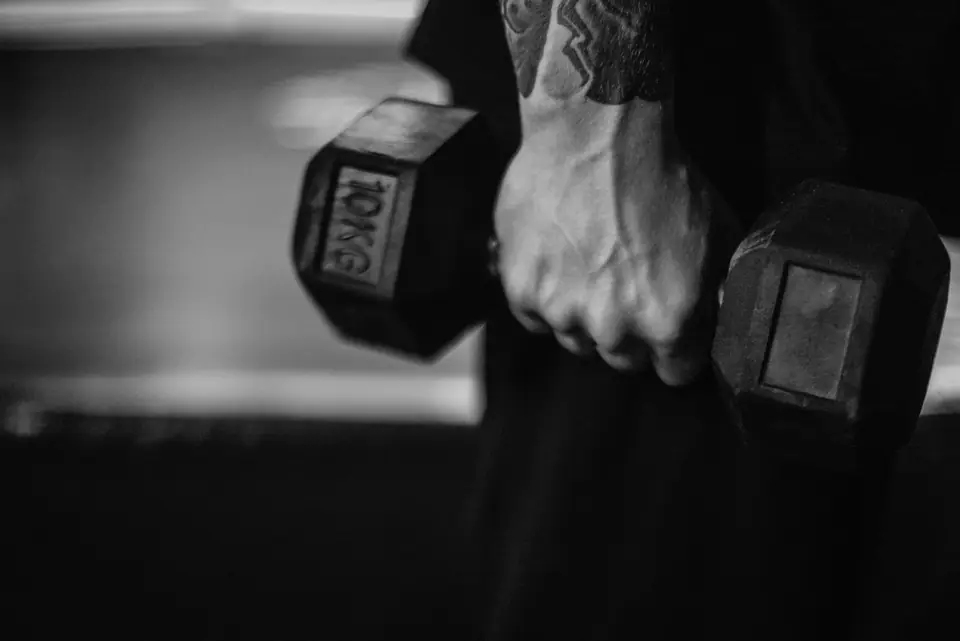 A tattooed hand holds a hexagonal dumbbell marked 20 kg against a dark, blurred background.