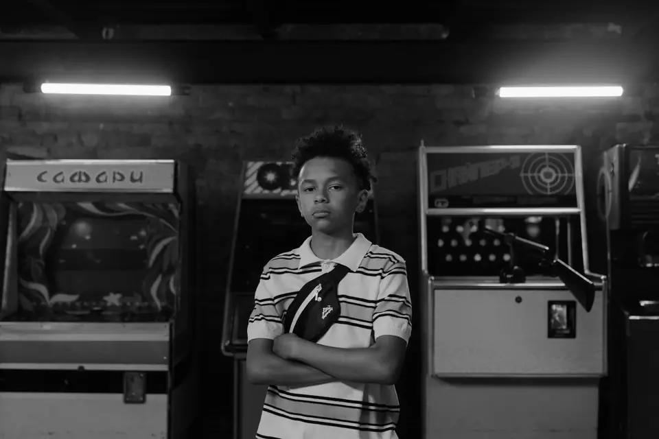 A boy in a striped shirt stands with folded arms between two vintage arcade game cabinets.