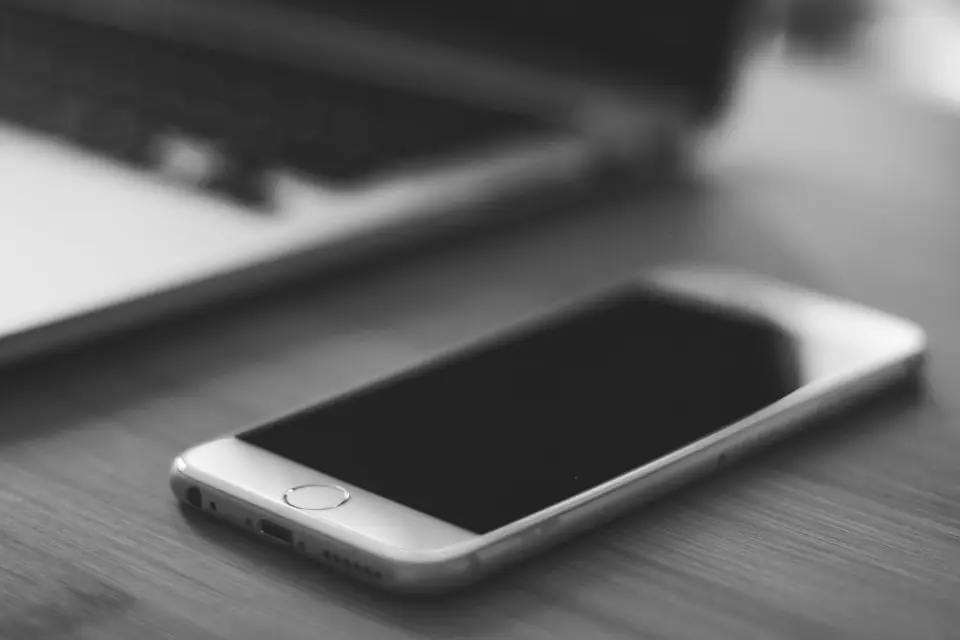 A silver smartphone with a dark screen lies on a desk in front of a blurred laptop.