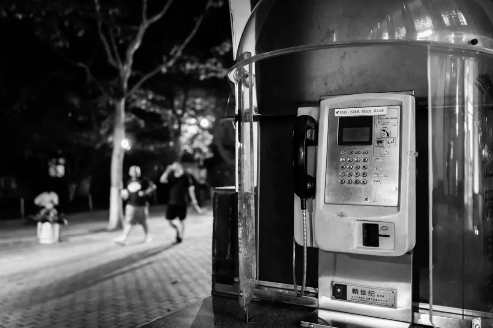 A public pay phone with a keypad stands in a metal kiosk on a lit sidewalk at night.