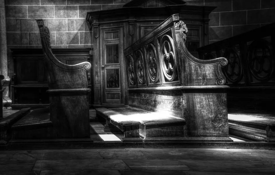 Ornately carved wooden choir stalls stand against a stone wall in a dim chapel.