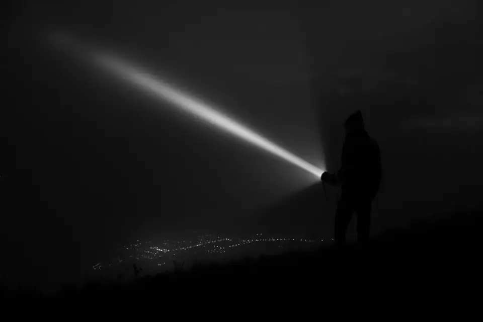 A silhouetted person on a dark hill shines a bright flashlight beam into misty night air.