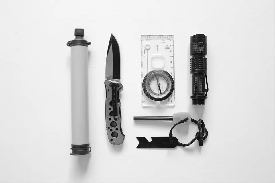 A water filter straw, a folding knife, a compass, a flashlight and a fire starter lie in a row on white.