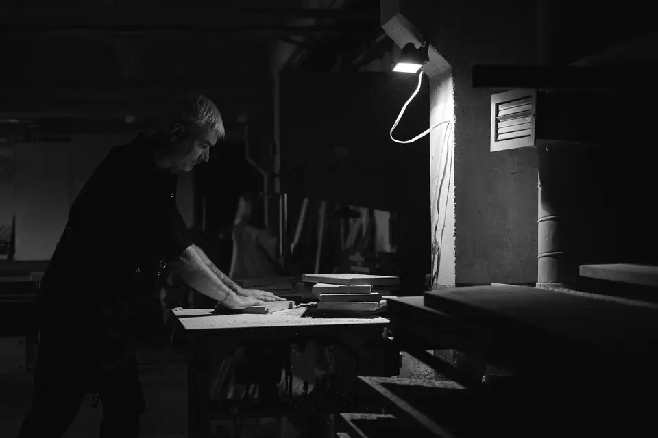 A man leans over a workbench in a dark woodshop, lit by a single hanging lamp.