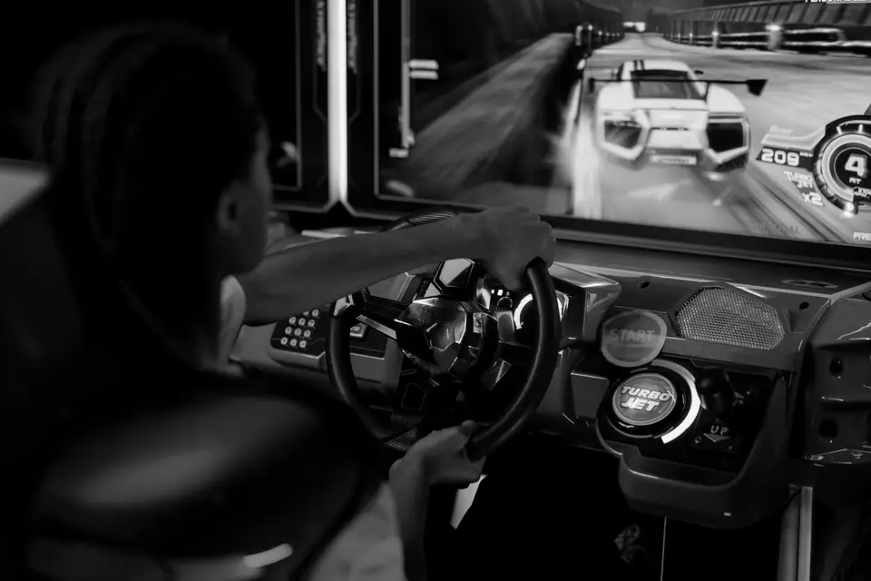 A person grips the steering wheel of an arcade driving game with a race car on screen.