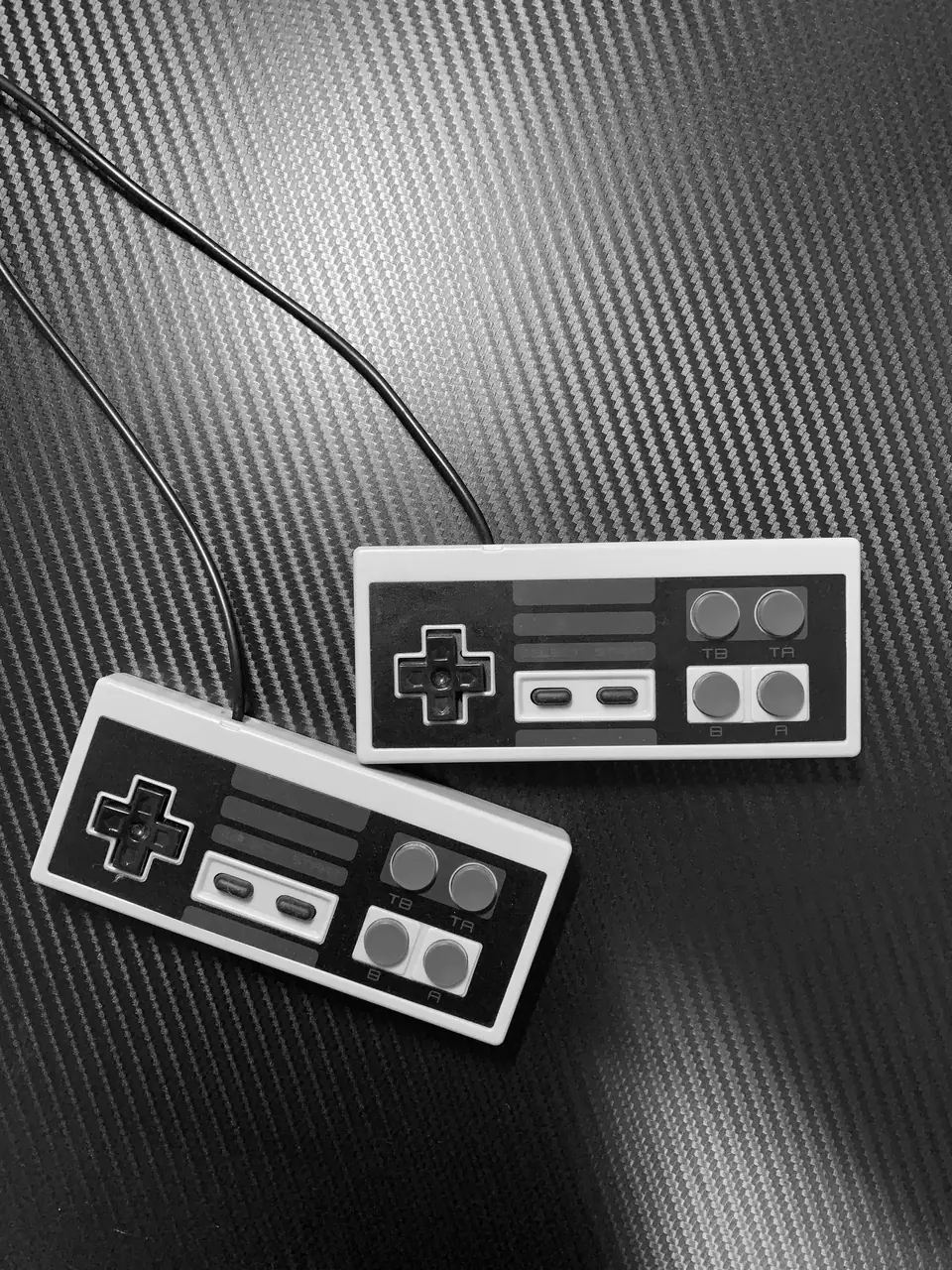 Two rectangular retro game controllers with cables lie on a dark carbon-fiber surface.