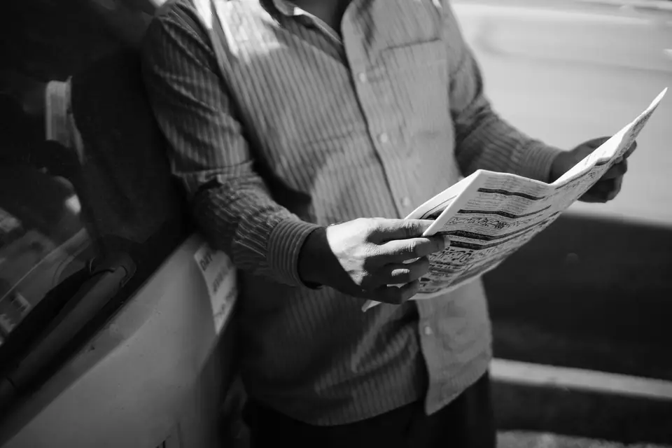 A man in a striped shirt leans against a car and reads an open newspaper held in both hands.
