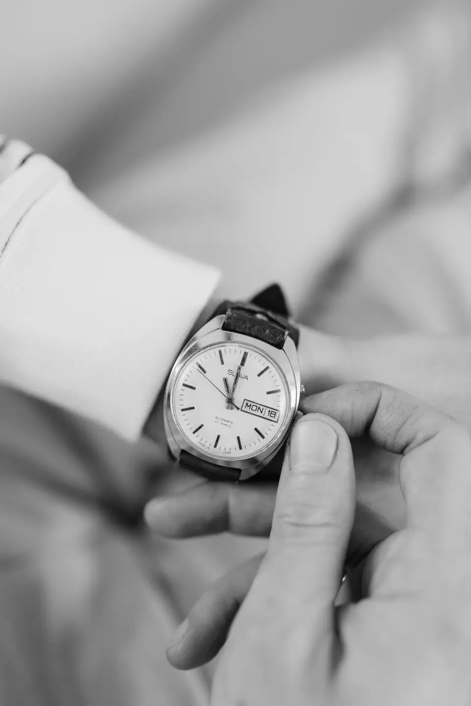 A hand adjusts the crown of a wristwatch with a white dial and day-date window on a shirt-cuffed wrist.