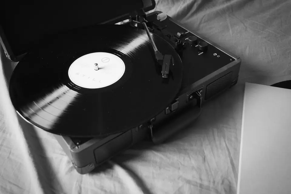 An open portable turntable with a vinyl record on its platter rests on wrinkled bed linen.