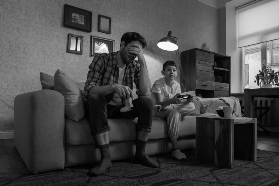 A man holds his hand over his face on a sofa while a boy beside him plays a video game.