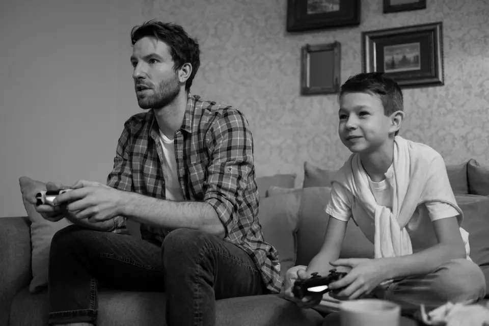 A bearded man and a young boy sit on a sofa, both playing a video game with controllers.