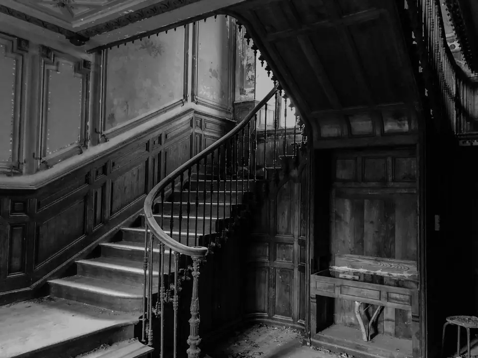A grand wooden staircase with an ornate railing rises through a faded, paneled hall.