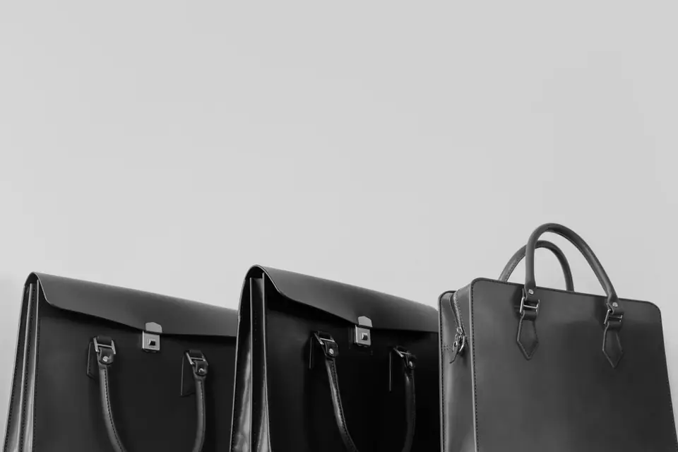 Three black leather briefcases and bags with buckles and top handles stand side by side against a pale wall.