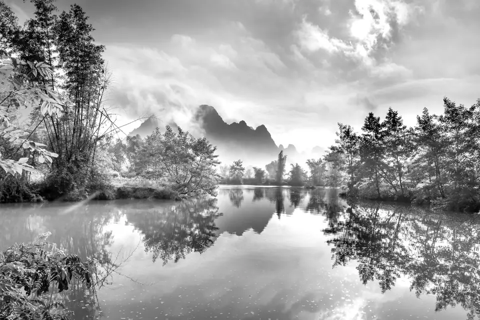 Still water reflects trees and steep distant peaks beneath a cloudy sky.