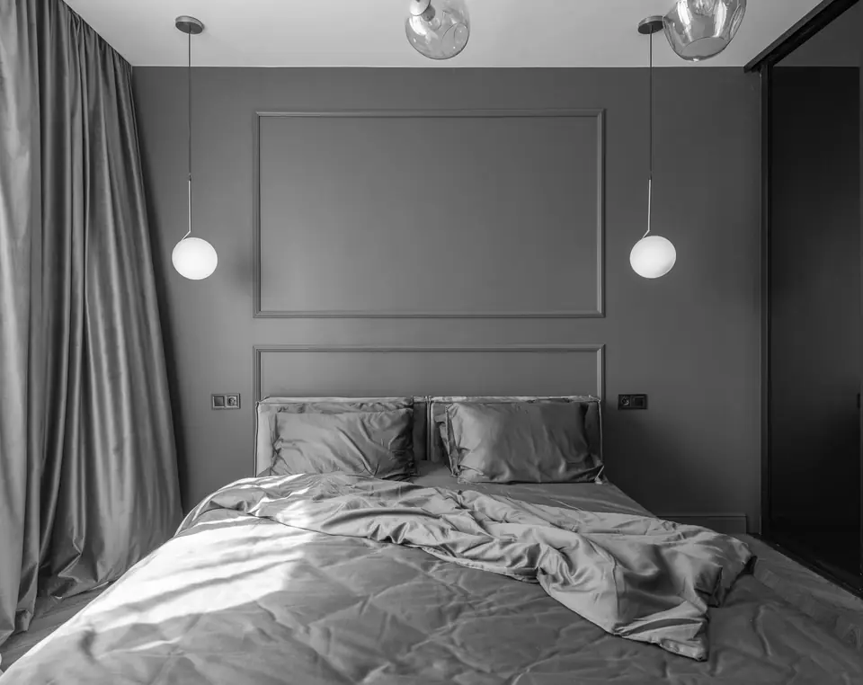 An unmade double bed with rumpled bedding stands between two hanging globe lamps and long curtains.