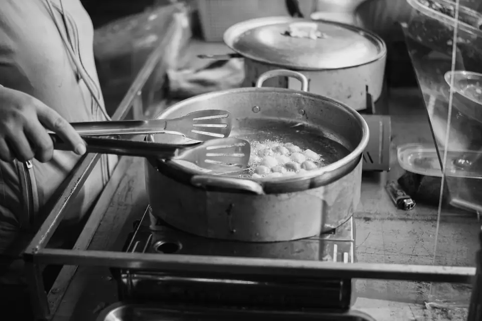 A cook uses tongs and a slotted spoon over a large steel pot on a stove in a busy kitchen.
