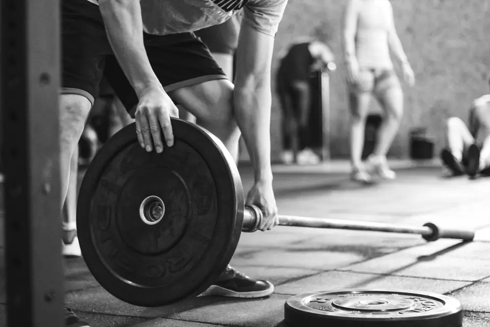 A person loads a black weight plate onto a barbell, with other people exercising in the background.
