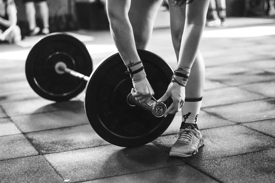 A person bends to fasten a clip on a barbell loaded with black weight plates on a gym floor.