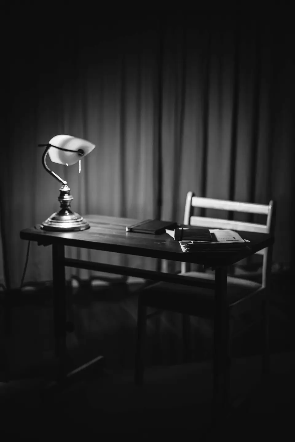 A small wooden desk with a banker’s lamp, a notebook and a chair stands in a dim room with curtains.