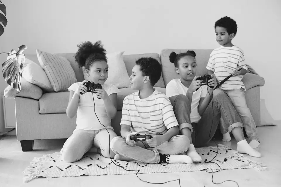 Four children sit on a rug in front of a sofa, several of them holding game controllers.