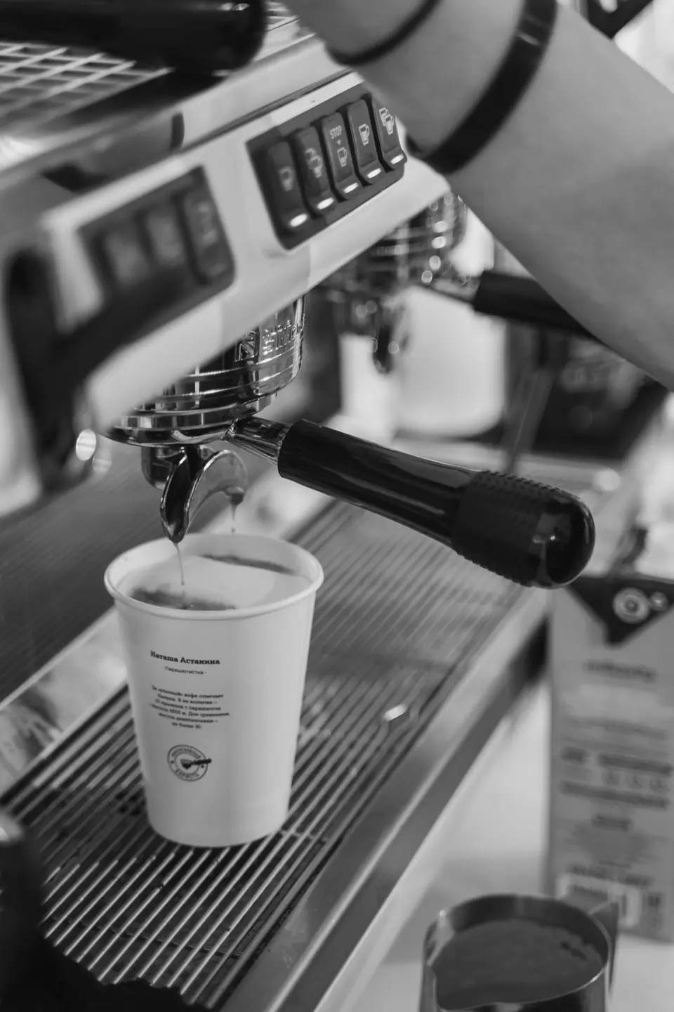 An espresso machine pours coffee from a portafilter into a white paper cup on a drip tray.