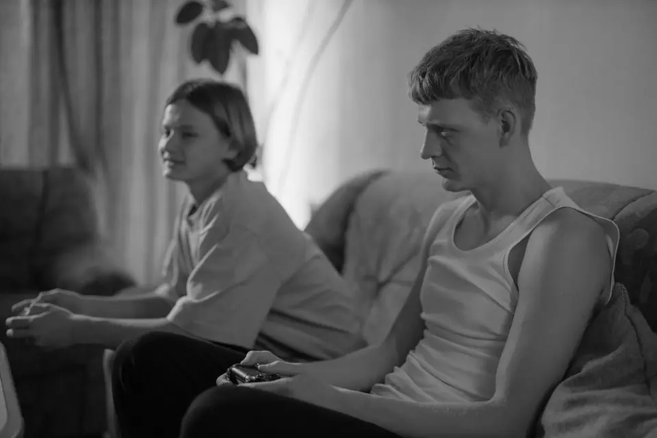 Two young men sit on a sofa holding game controllers and looking at an unseen screen.