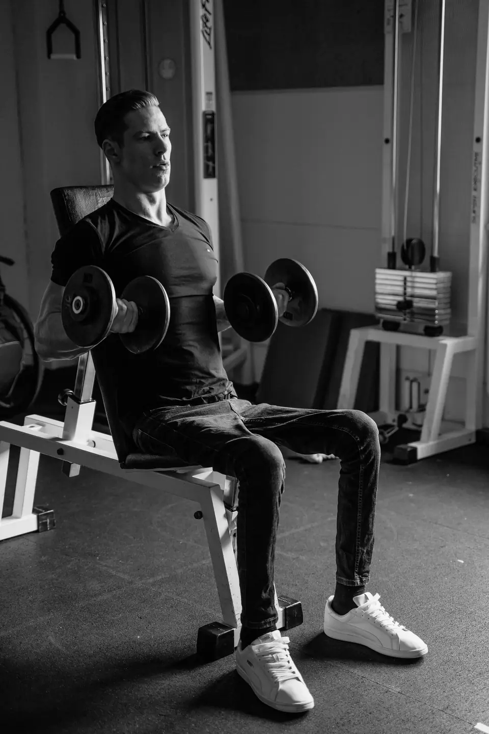 A man seated on a gym bench curls a pair of dumbbells in a room with cable machines.