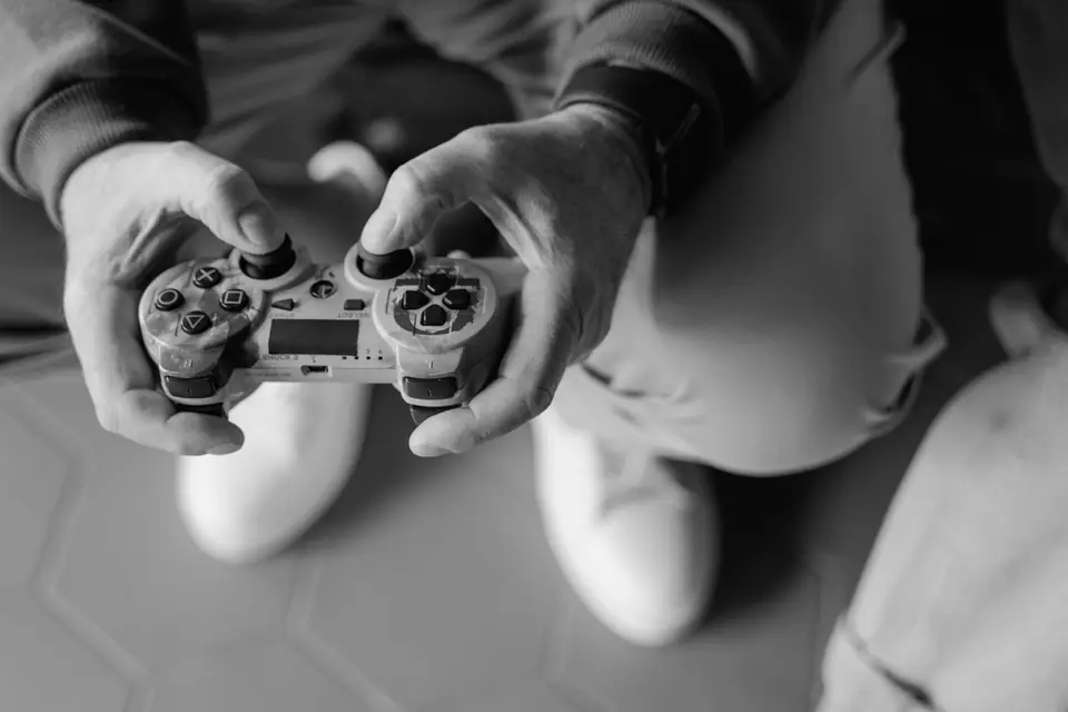 Two hands hold a modern game controller in front of a seated person’s knees and white sneakers.