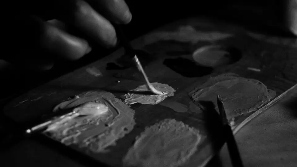 A hand holds a brush over a palette covered with thick daubs of mixed paint.