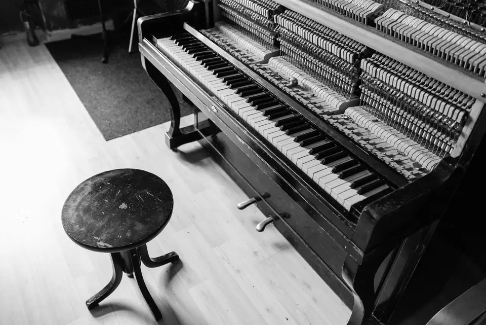 An upright piano with its front panel removed shows hammers and strings beside a round stool.