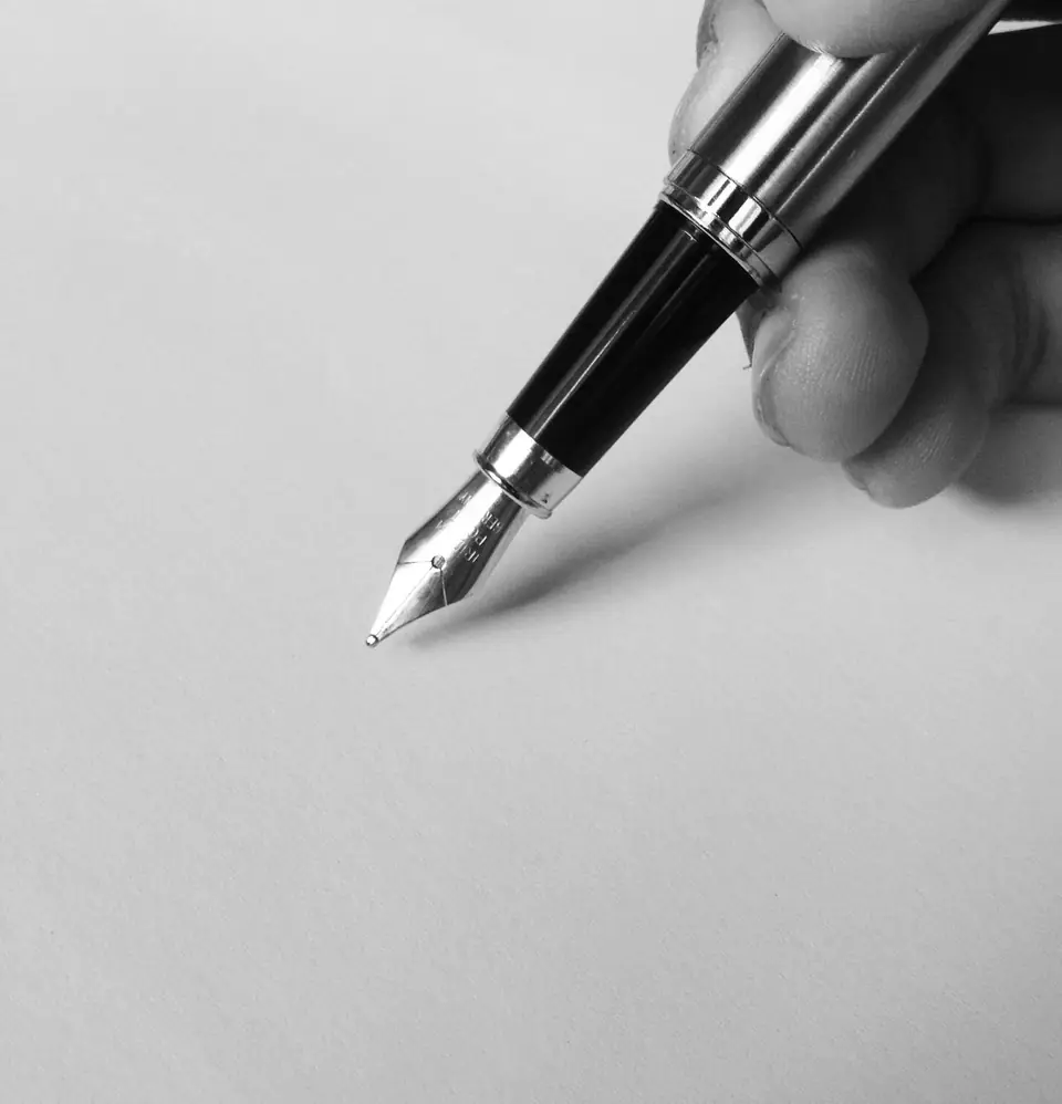 A hand holds a fountain pen with its nib just above a blank sheet of white paper.
