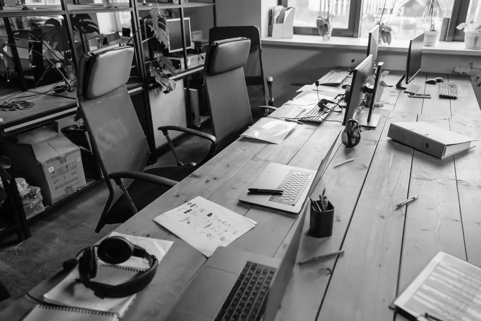 An open office has long wooden desks covered with monitors, laptops, headphones and papers near a bright window.