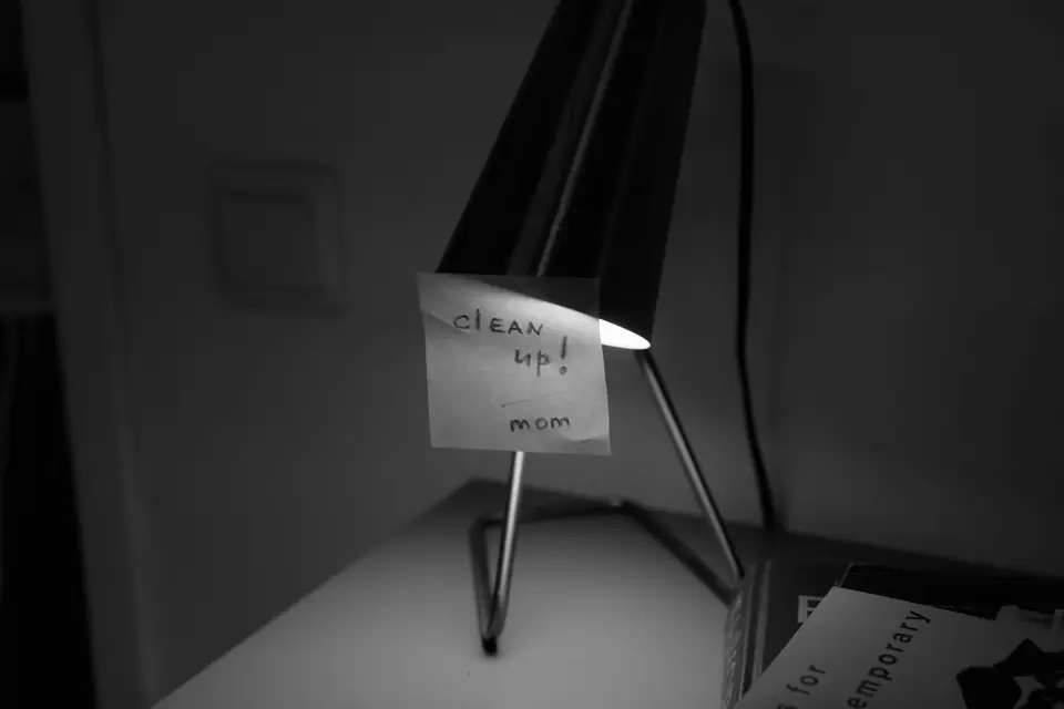 A handwritten note reading “clean up! mom” is taped to a desk lamp in a dim room.