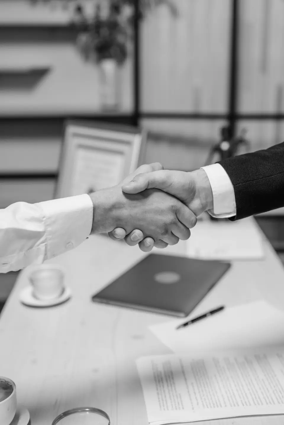 Two people shake hands across a desk holding documents, a pen, a folder and a coffee cup.