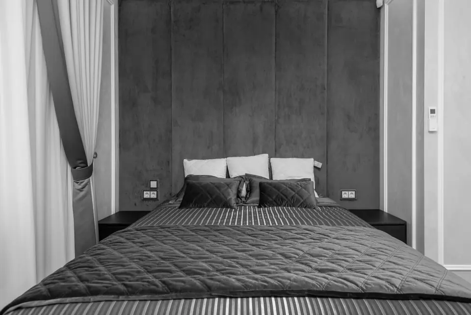 A neatly made double bed with dark bedding and pillows stands against a tall padded headboard.