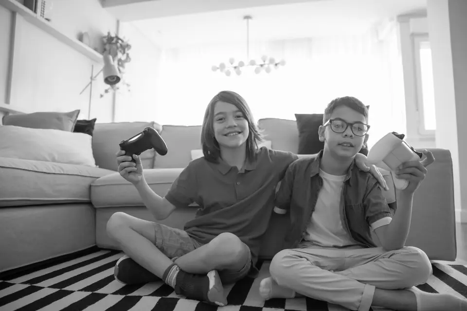 Two smiling boys sit cross-legged on a striped rug, one holding a game controller.