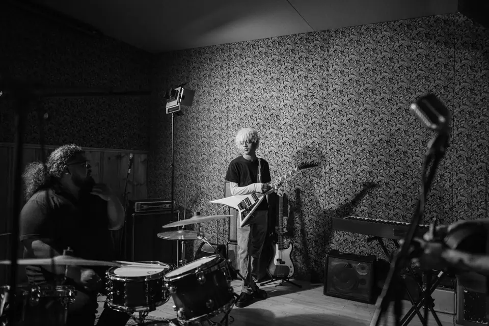 A drummer and a guitarist rehearse in a small room with patterned wallpaper.