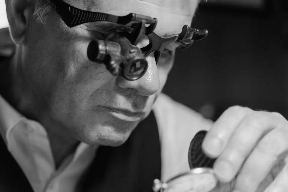 A gray-haired man wearing binocular magnifying loupes closely examines a pocket watch in his hand.